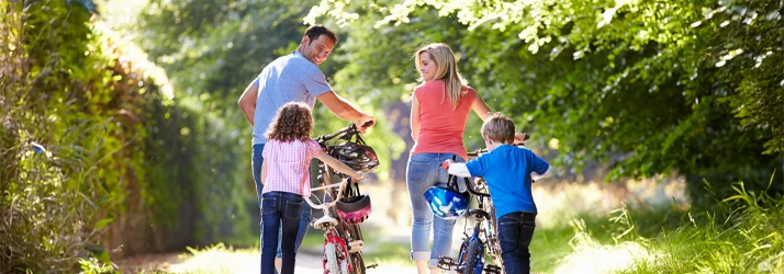 Four people walking with two bicycles along a tree-lined path outdoors.